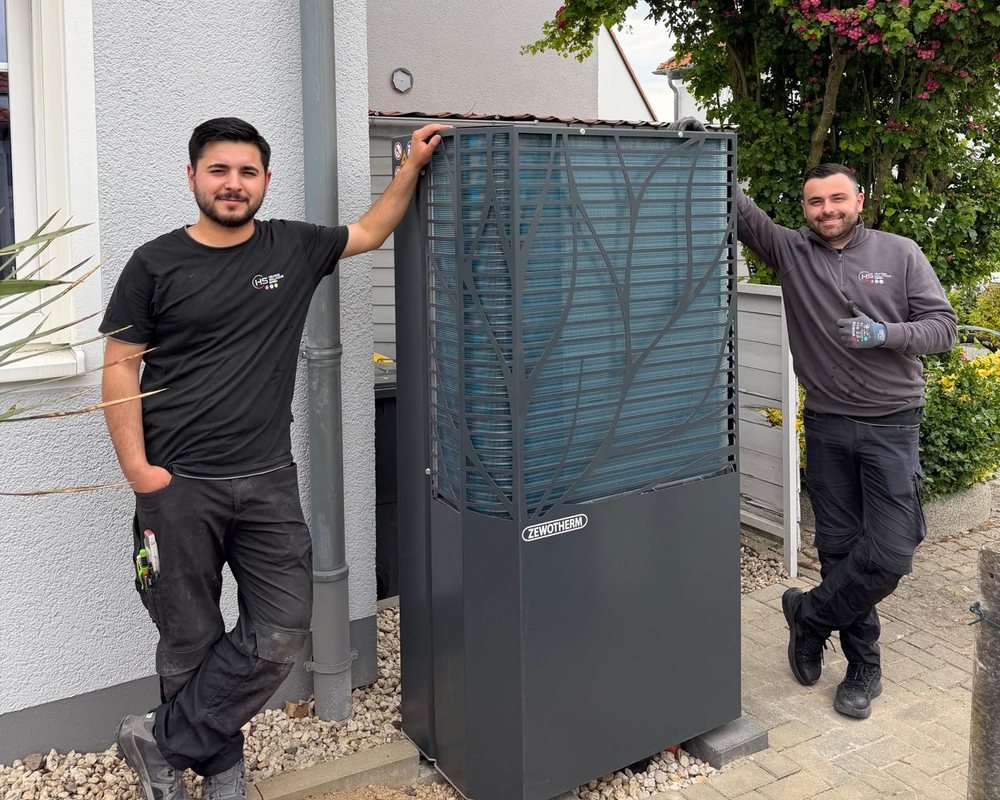 Zwei Monteure von HS Heating Solutions neben einer Wärmepumpen-Außeneinheit an einem Wohnhaus