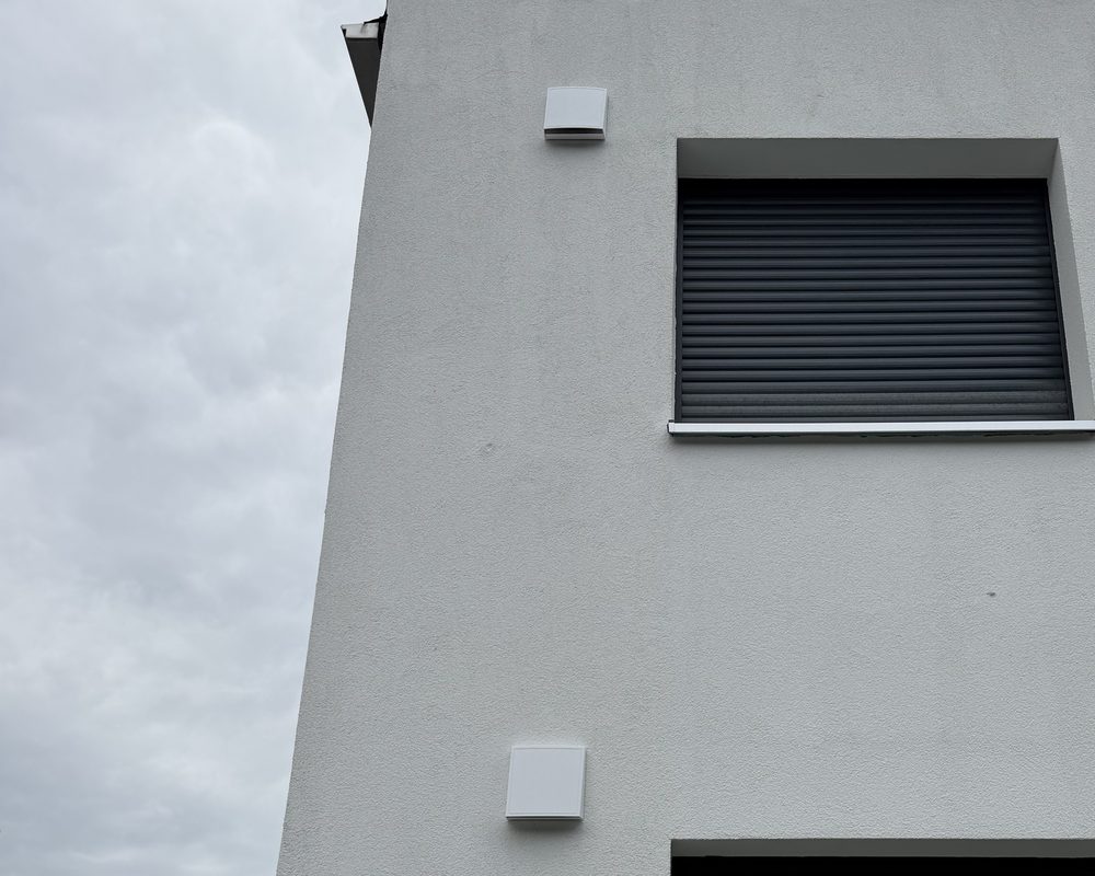 Fassade eines Wohnhauses mit zwei weißen Außenhauben neben einem Fenster mit Rollladen